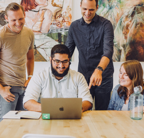 2 people standing and 2 people sitting at a timber table. One person sitting as a laptop in front of them.