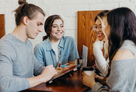 Four people sitting around a timber table. There is a laptop and an iPad. One person is holding a coffee cup.