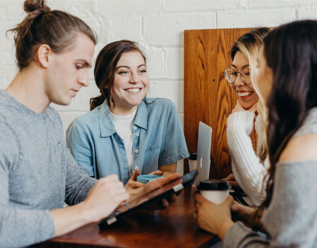 Four people sitting at a table with a laptop and iPad. One person is holding a coffee