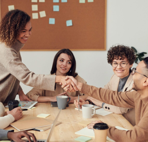 A group of people sitting around a timber table. Two people are shaking hands across the table. There are coffee mugs, books and paper on the table.