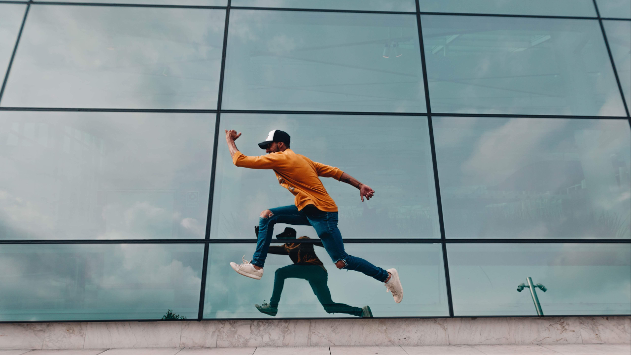 A male dressed in an orange top and blue jeans jumping in front of a glass building