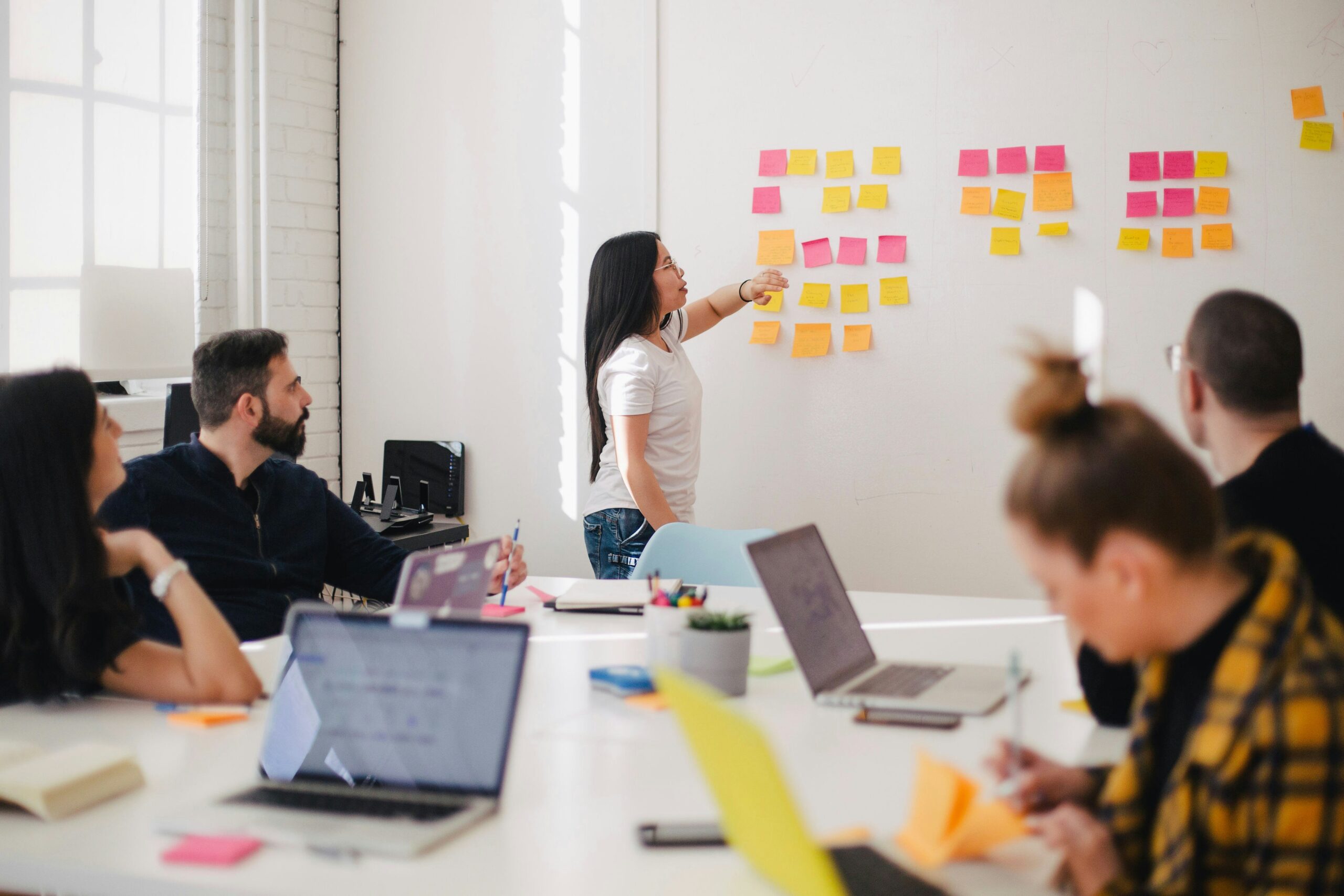 A group of people sitting around a large white desk in an office listening to a female standing by a whiteboard covered in post-it notes