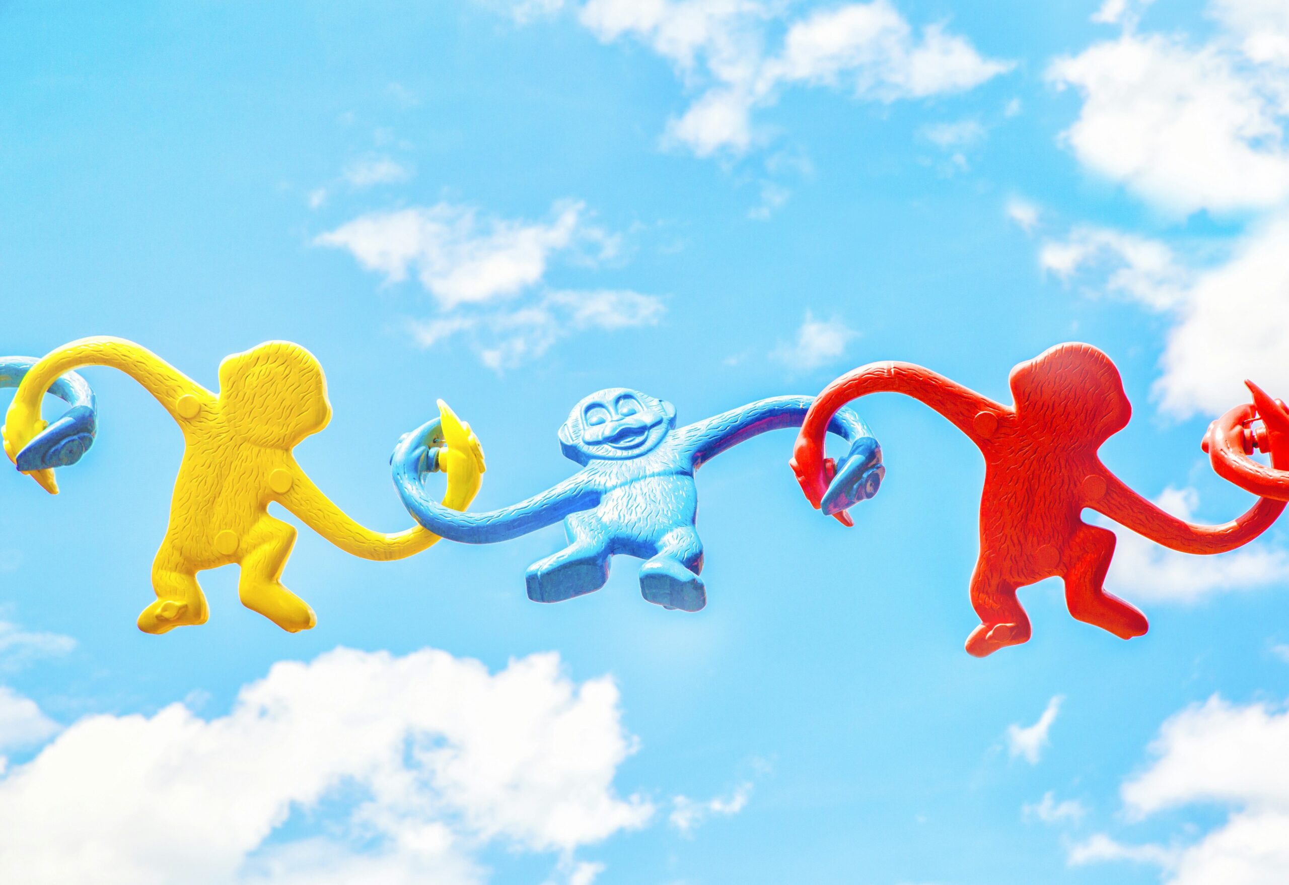 Three toys monkeys coloured yellow, blue and red linking arms with blue sky and white clouds behind them