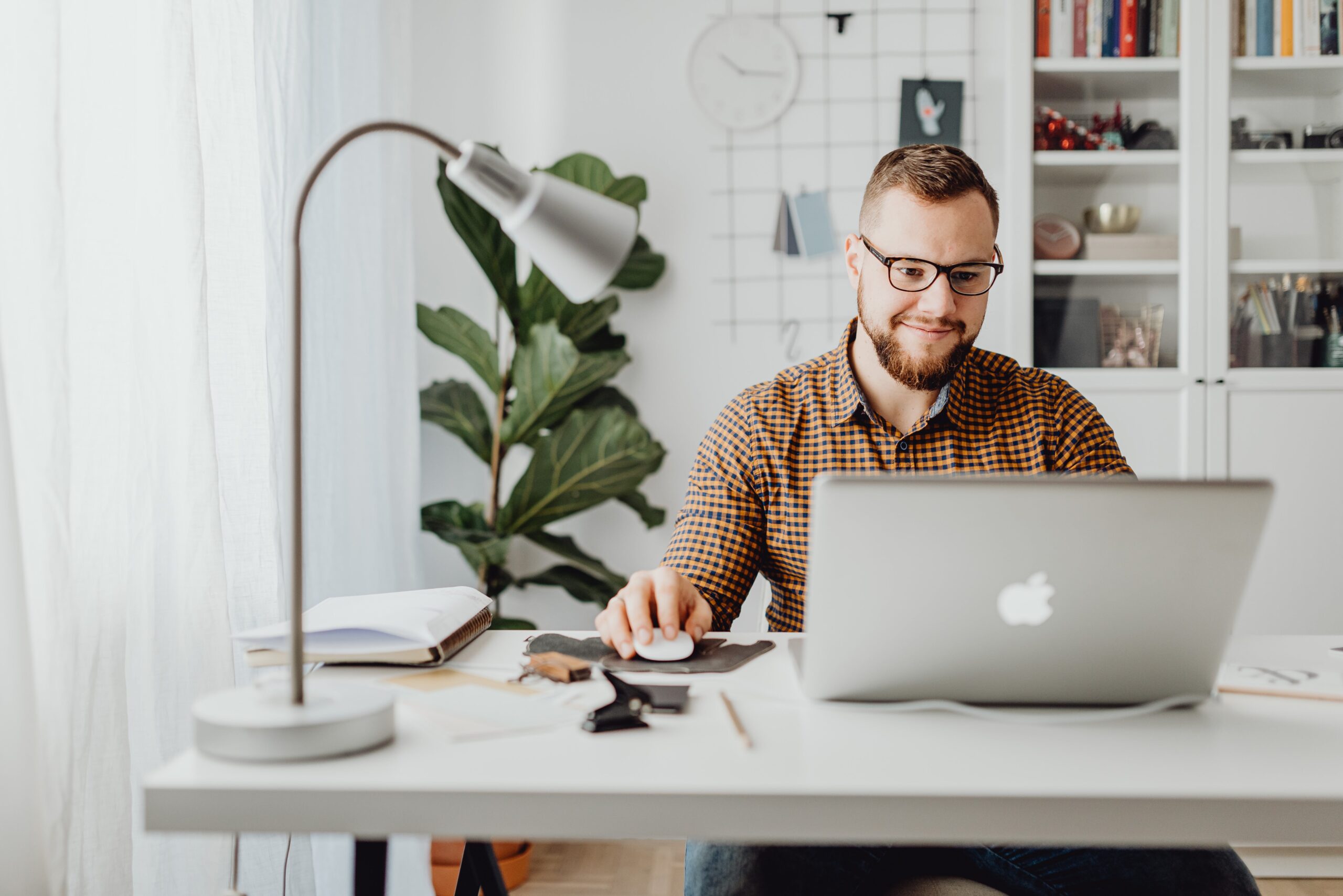 a man sitting at a white desk in his home office. He is looking at a laptop, there is a lamp on his desk