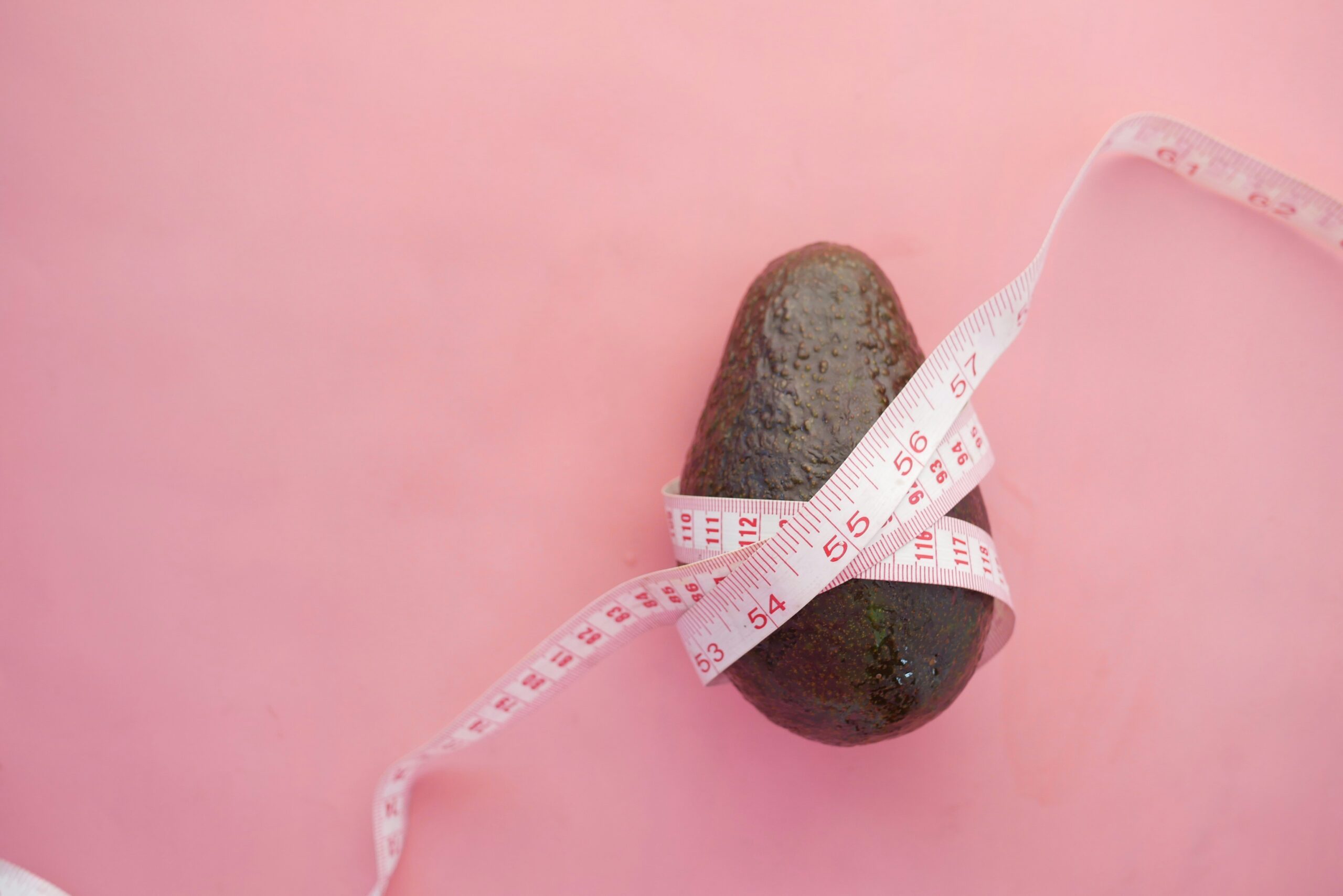 An avocado wrapped in a tape measure on a pink background